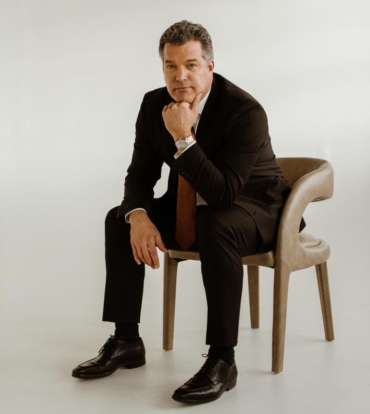 A man in a black suit sits on a beige chair, resting his chin on his hand and looking at the camera against a plain light background.