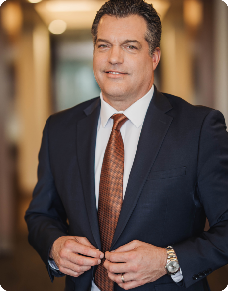 A man in a dark suit and brown tie stands indoors, buttoning his jacket and looking at the camera. The background is softly blurred.