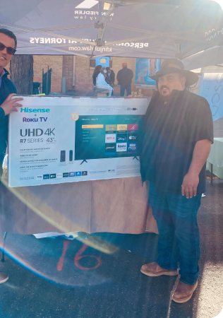 Two men stand under a tent, holding a large boxed Hisense Roku TV, UHD 4K 43" R7 Series, at an outdoor event.