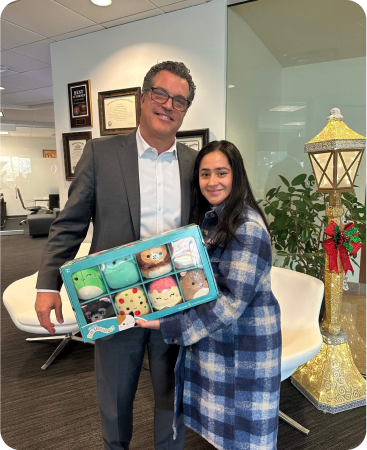 A man in a suit and a woman in a plaid coat stand indoors holding a box of plush toys. Office furniture, framed certificates, and a holiday lamp decoration are visible in the background.
