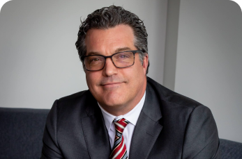 A man with dark curly hair and glasses wearing a dark suit, white shirt, and red striped tie sits on a couch in an office setting.