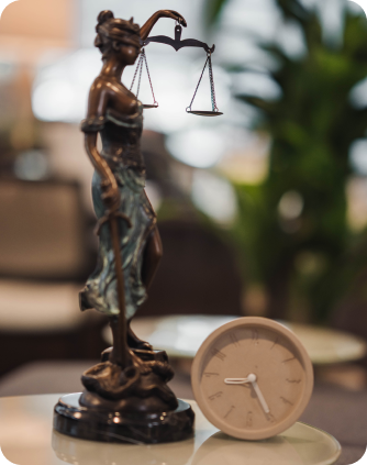 A bronze statue of Lady Justice holding scales stands next to a small round clock on a table, with a blurred background.