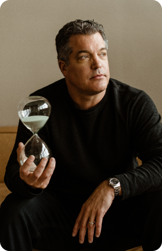 A man in a dark sweater sits holding an hourglass in his right hand and resting his left hand on his knee, looking slightly to the side.