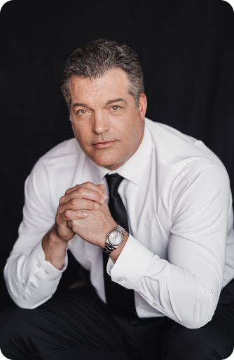 A man in a white dress shirt and black tie sits with his hands clasped, looking at the camera against a dark background.