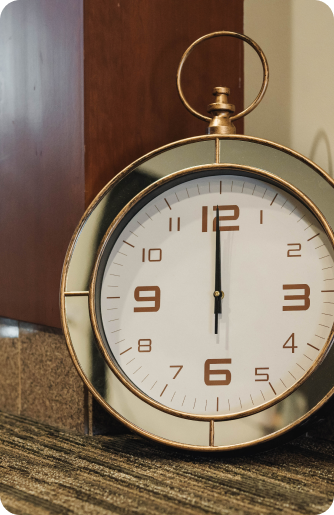 A round, gold-rimmed clock showing the time as 12:00, resting against a wooden surface on a carpeted floor.