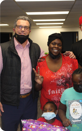 A man and a woman stand together indoors, smiling at the camera. Two children wearing masks are in front of them, one holding a wrapped gift.