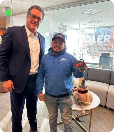 Two men stand indoors near a table; one is dressed in a suit and the other in casual clothes, holding a small object. A dog wearing a sweater sits on the table.