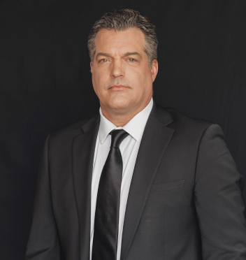 A middle-aged man in a black suit, white shirt, and black tie poses against a plain dark background.
