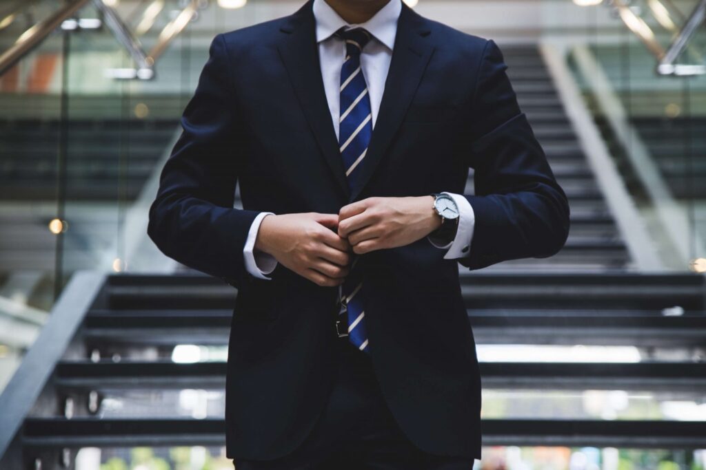 A person in a dark suit and striped tie adjusts their jacket while standing in front of a staircase in a modern building.