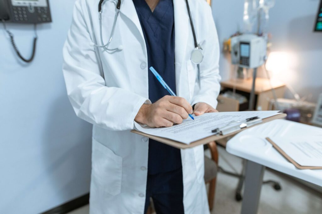 A doctor in a white coat and stethoscope writes on a clipboard in a medical office.