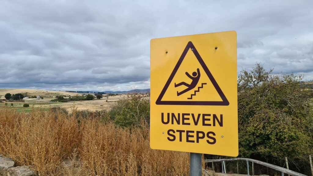 Yellow warning sign with a graphic of a person tripping on stairs and the text "UNEVEN STEPS" in a rural outdoor area.