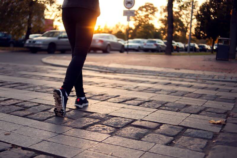 Person walking across a cobblestone street at sunset with parked cars and trees in the background.
