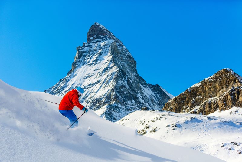 Person skiing downhill on fresh snow with the Matterhorn mountain in the background under a clear blue sky.