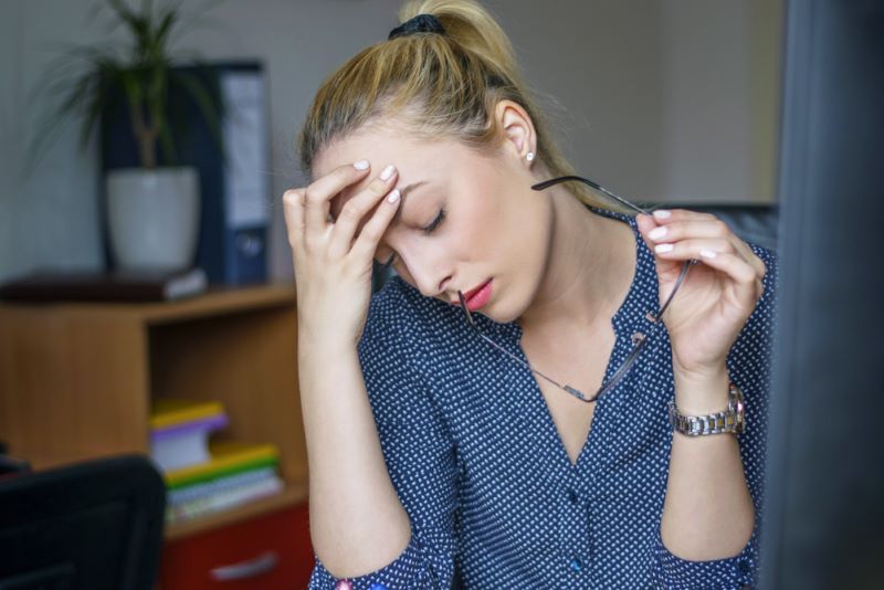 A woman sits at a desk holding her glasses and resting her head in her hand, appearing stressed or tired.