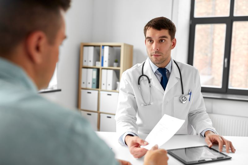 A doctor in a white coat hands a paper to a patient across a desk during a consultation in a medical office.