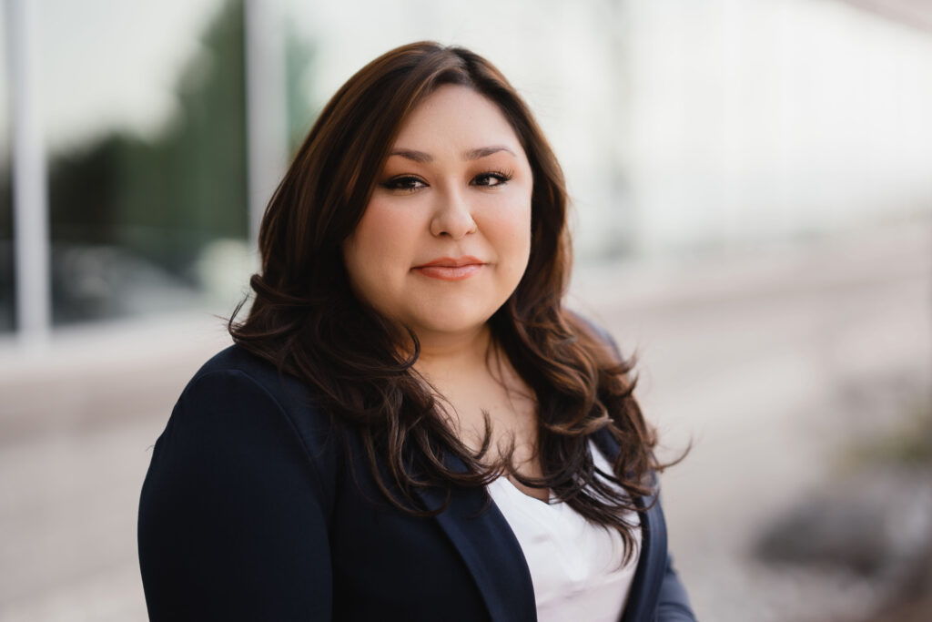 A woman with long dark hair, wearing a navy blazer and white top, poses outdoors in front of a blurred building.