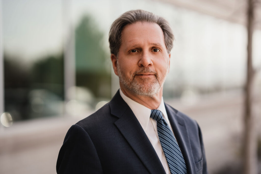 Middle-aged man with gray hair and beard, wearing a dark suit, white shirt, and blue striped tie, standing outdoors in front of a blurred glass building.