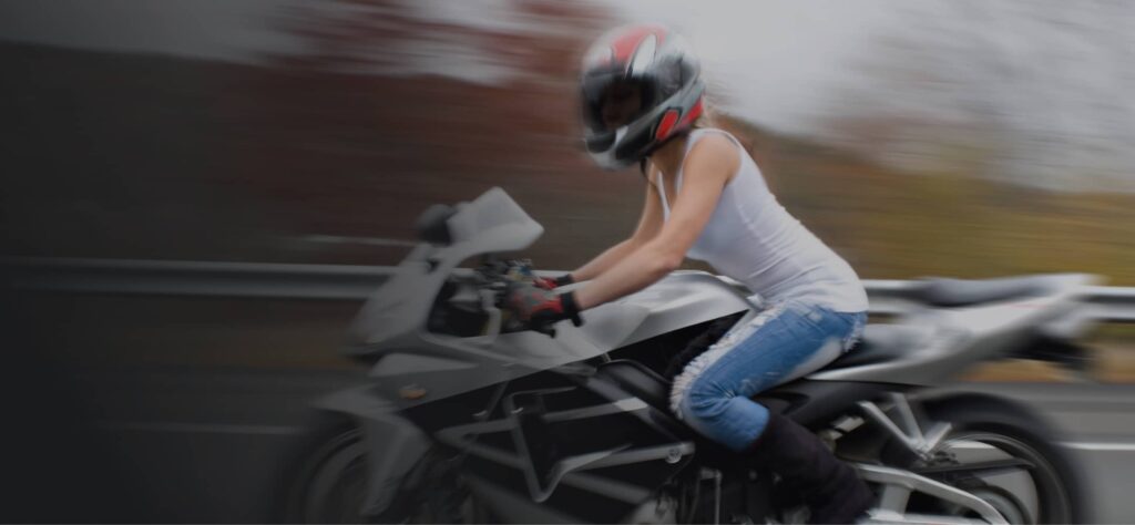 A person wearing a helmet, white tank top, and jeans is riding a black sports motorcycle on a road with a blurred background.