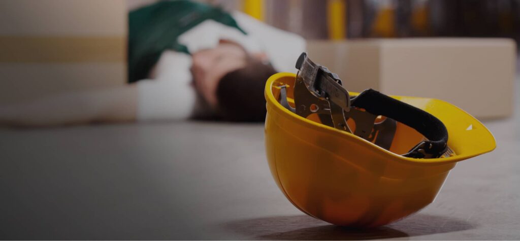 A yellow hard hat lies on the floor in the foreground, while a person is lying down in the background near cardboard boxes.