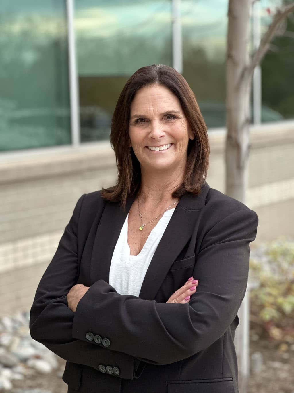 Woman with shoulder-length brown hair wearing a black blazer and white blouse, standing outdoors with arms crossed, smiling at the camera.