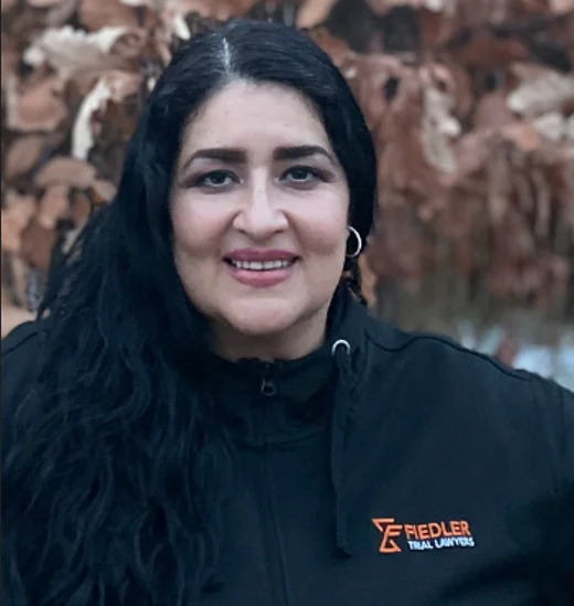 A woman with long black hair smiles while wearing a black Fiedler Trial Lawyers jacket, standing outdoors with brown leaves in the background.