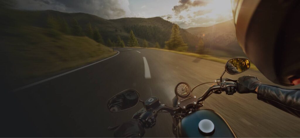 A person rides a motorcycle on a winding mountain road at sunset, with trees and hills visible in the background.