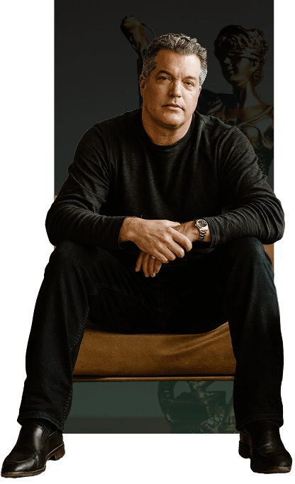 A middle-aged man in a dark sweater and jeans sits on a chair with his hands clasped, in front of a classical sculpture background.