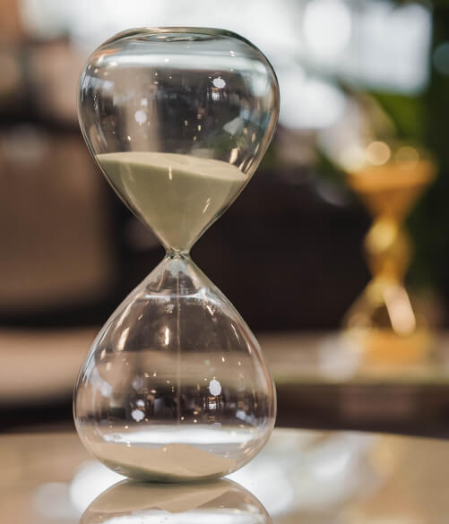 A glass hourglass with white sand partially fallen to the bottom sits on a reflective surface; a blurred gold hourglass is in the background.