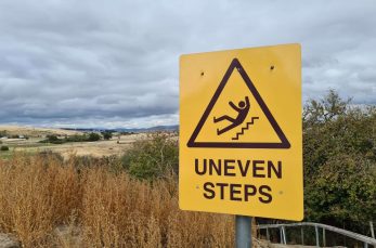 Yellow warning sign with a graphic of a person tripping on stairs and the text "UNEVEN STEPS" in a rural outdoor area.