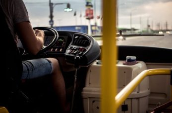 A person wearing shorts is driving a bus, with their hand on the steering wheel and dashboard controls visible. The view is from inside the bus, looking toward the front windshield.