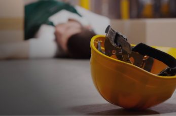 A yellow hard hat lies on the floor in the foreground, while a person is lying down in the background near cardboard boxes.