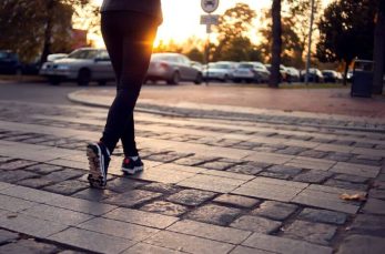 Person walking across a cobblestone street at sunset with parked cars and trees in the background.