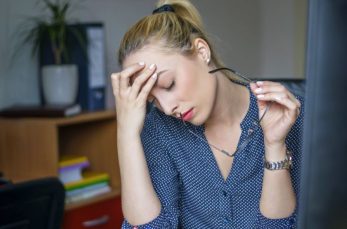 A woman sits at a desk holding her glasses and resting her head in her hand, appearing stressed or tired.